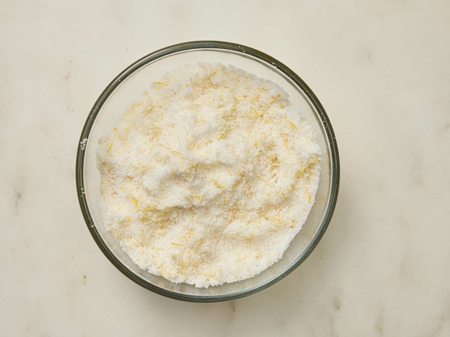 lemon zest and sugar combined together in a glass bowl on a marble surface 