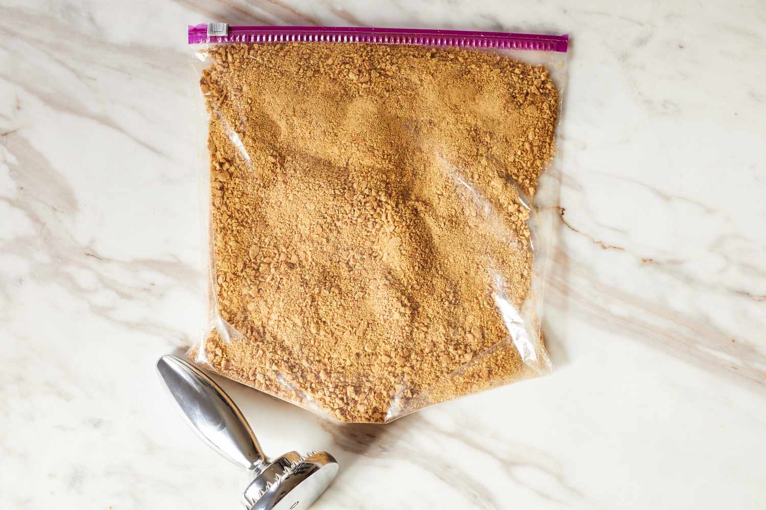 A zippered plastic bag filled with finely crushed crumbs on a marble surface with a metal tamper placed nearby