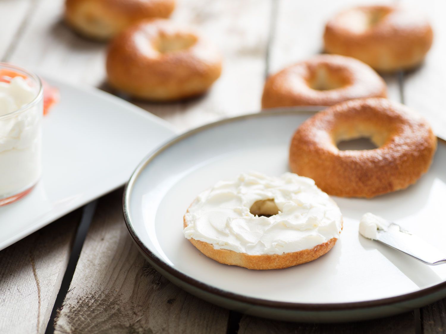 Split bagel on a white plate topped with cream cheese. Additional bagels and toppings in the background.
