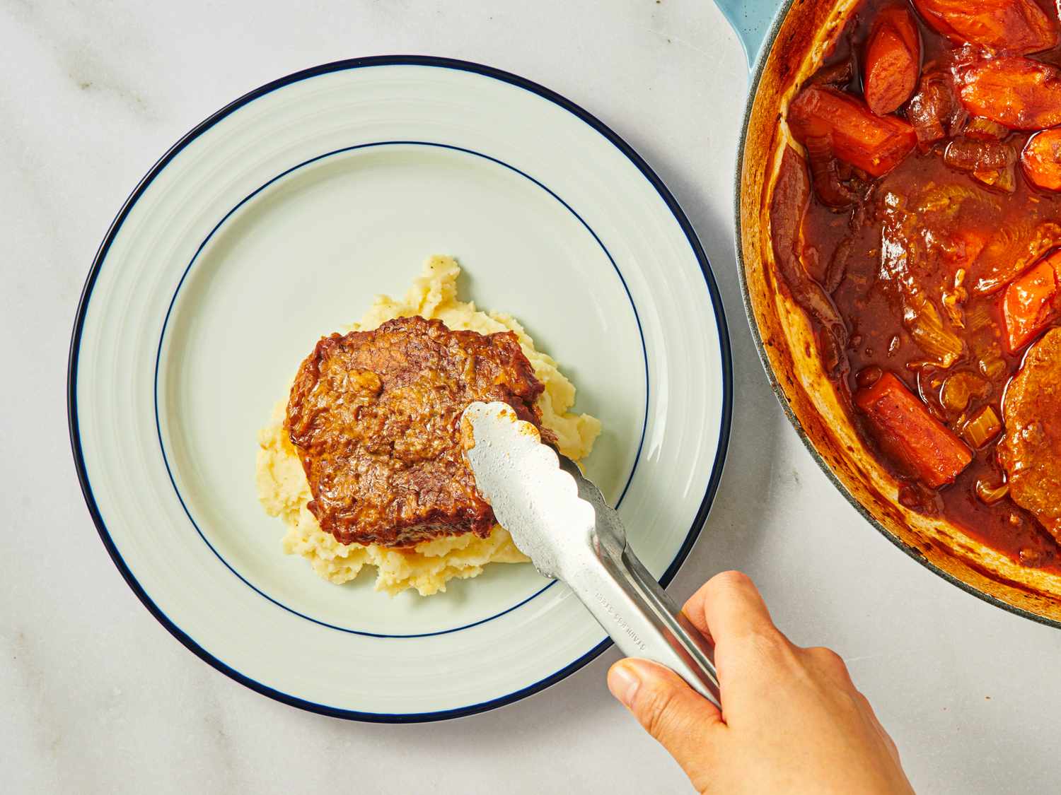 A person placing a cooked meat patty onto mashed potatoes using tongs next to a skillet containing a stew with carrots and broth