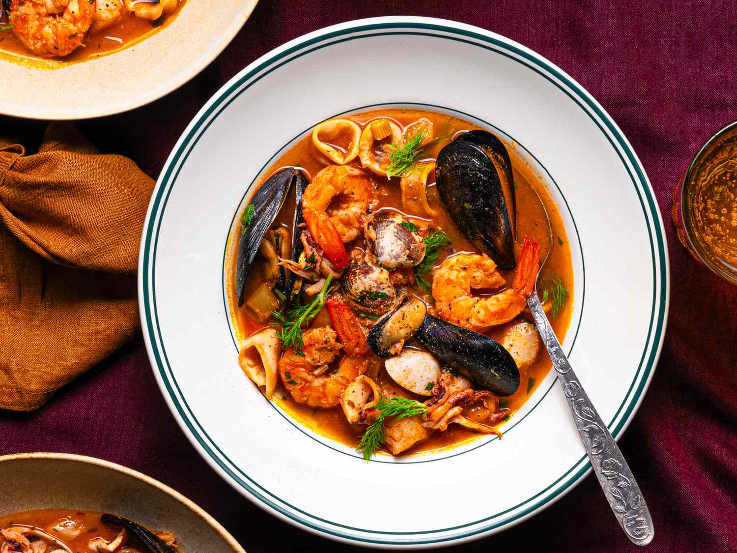 A seafood soup served in a white bowl with a spoon garnished with shrimp mussels and herbs placed on a maroon tablecloth