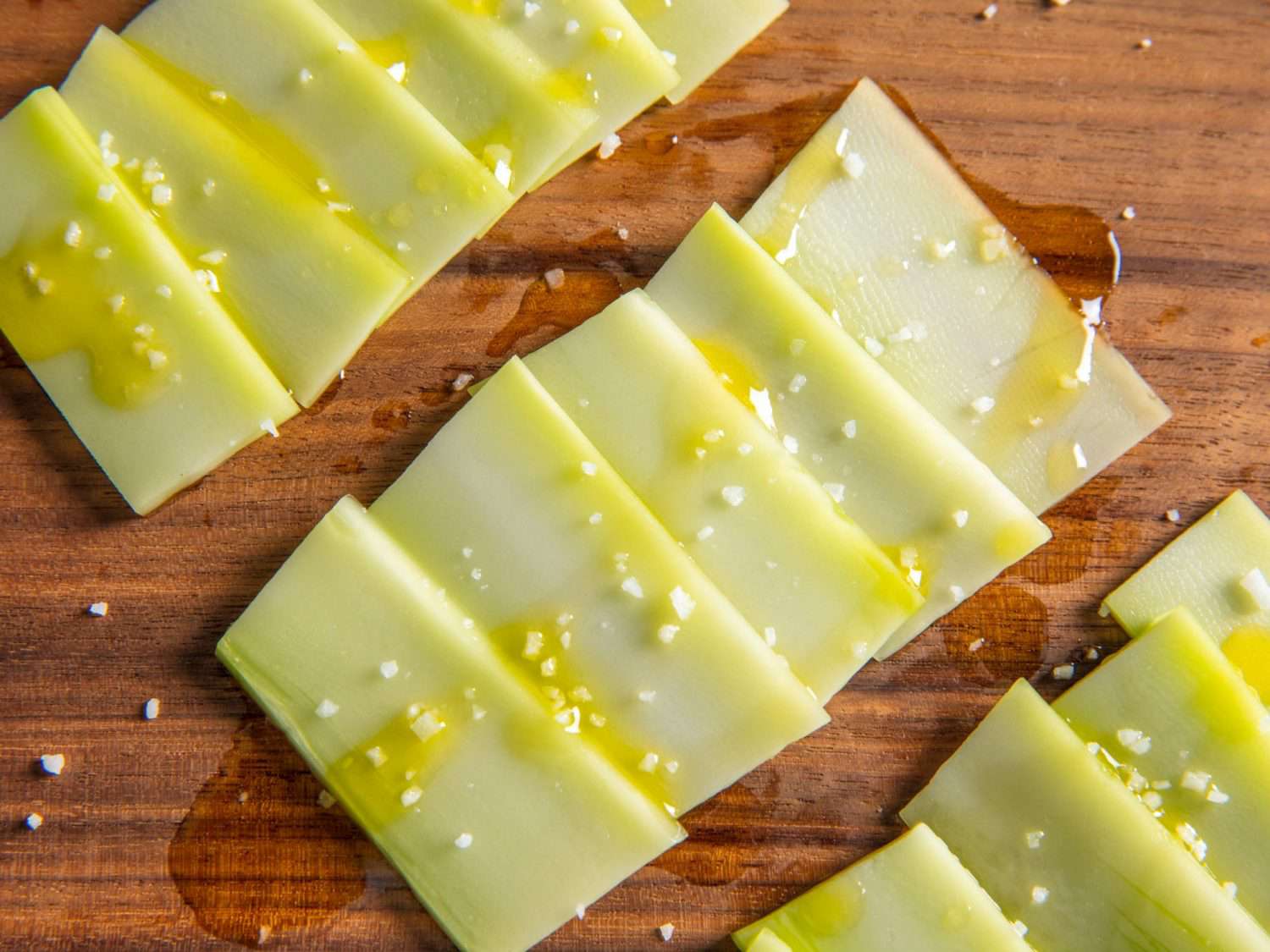 Thin planks of peeled broccoli stem, shingled on a wooden surface and topped with a drizzle of olive oil and a light sprinkle of flaky salt.