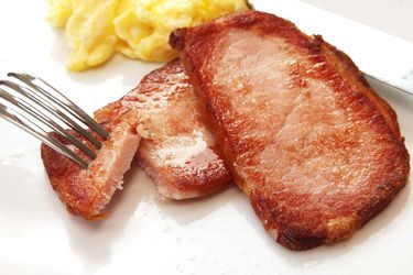 Closeup of browned slices of sous vide Canadian bacon, served on a white plate with scrambled eggs.