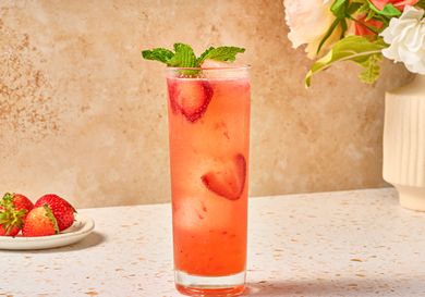 A strawberry mint sparkler served in a glass with mint garnish set on a table near flowers and fresh strawberries