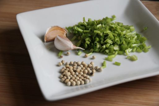 A square white ceramic plate holding white peppercorns, two unpeeled cloves of garlic, and chopped cilantro stems.