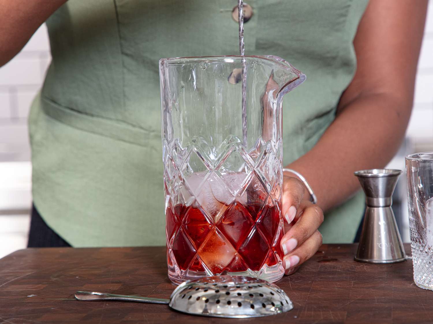 A person seen preparing a cocktail in a glass mixing vessel with ice a jigger nearby on the table