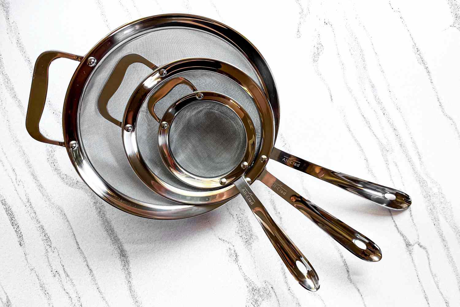 All-Clad Stainless-Steel Strainer, Set of 3 of varying sizes placed on a textured surface