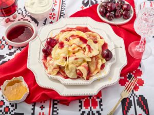 Cherry verenyky on decorative plates, on a printed surface, with a red napkin, and sides of cherry sauce, cream, and honey 