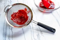 OXO Good Grips 8-Inch Double Rod Strainer with pressed fruit pulp beside a bowl containing a red liquid and a spatula