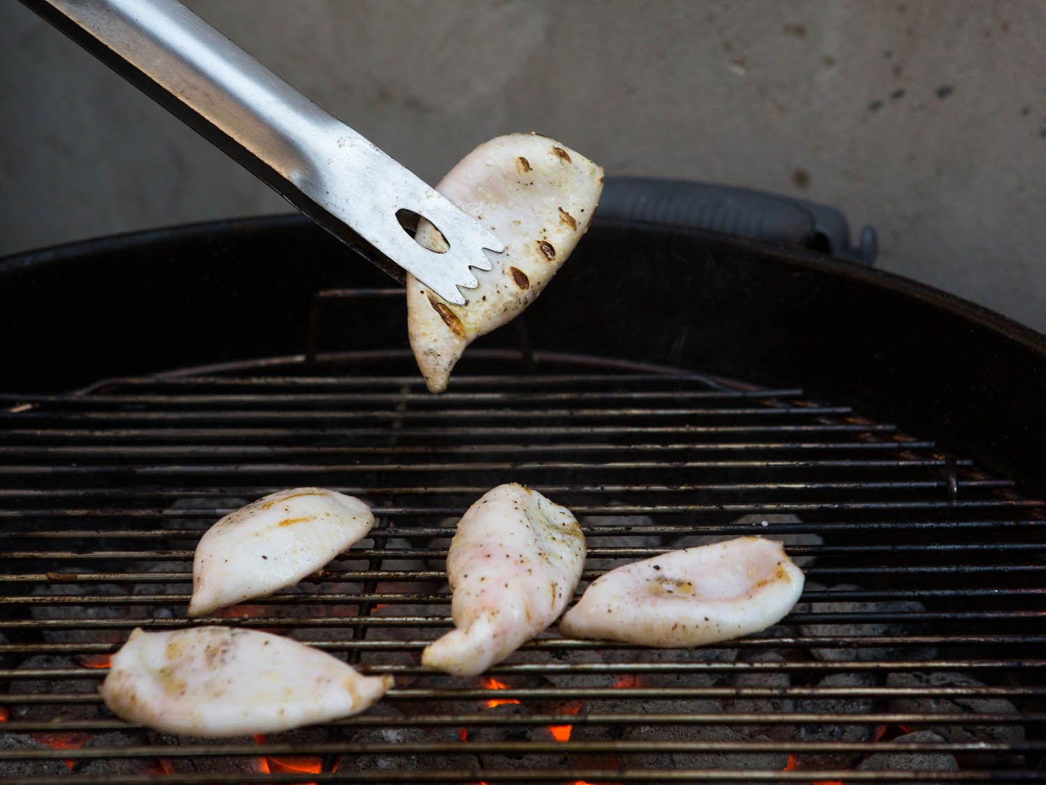 A pair of tongs is holding one of the charred squid bodes above the grill as the rest of them continue to cook.
