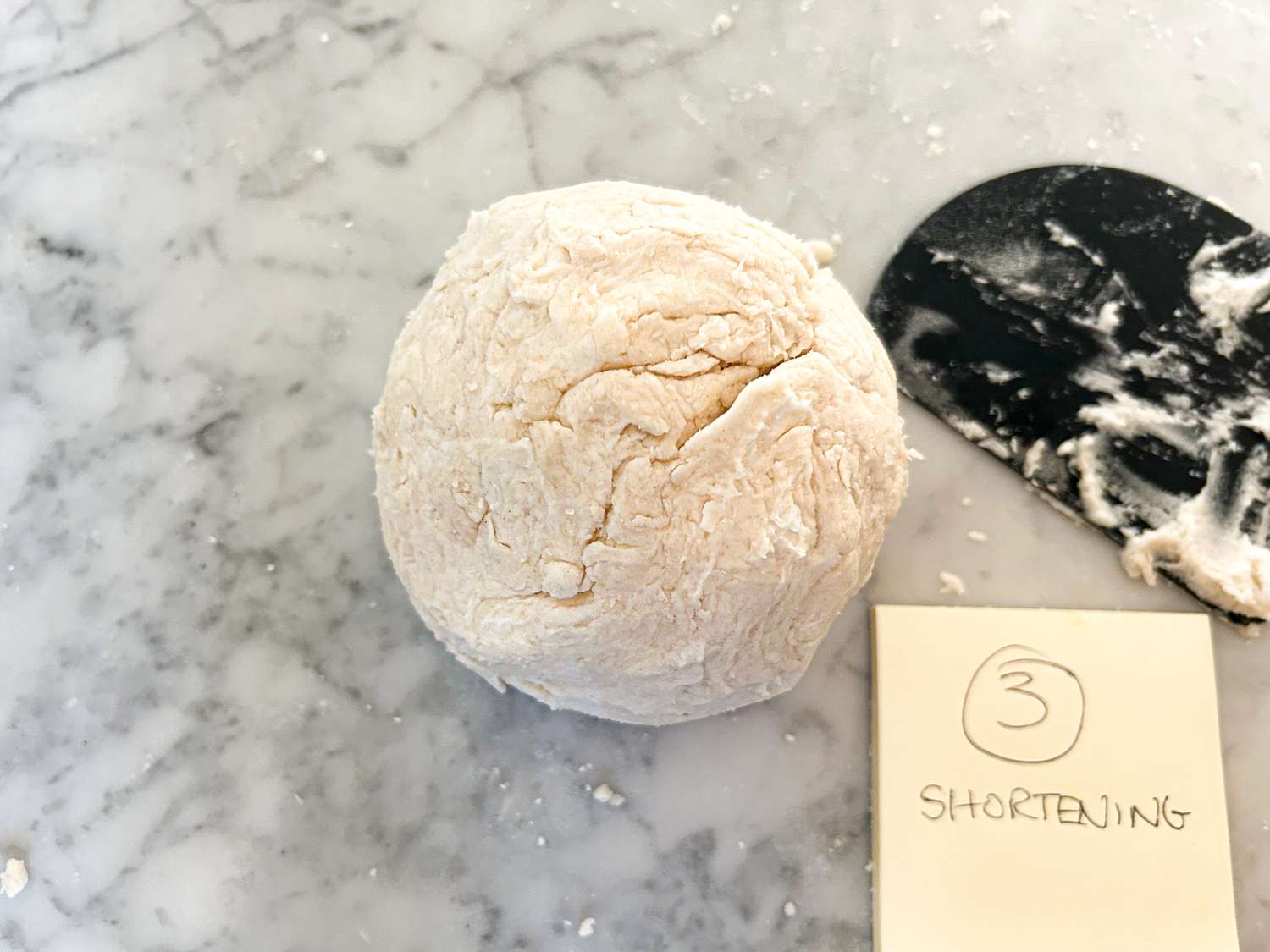 Ball of dough on a marble surface with a scraper and a note labeled Shortening 3 nearby