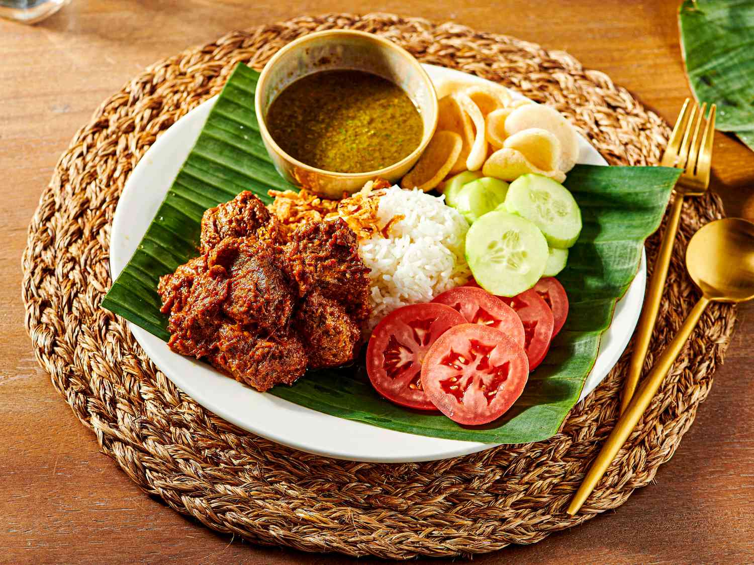 Plate of beef Rendang with rice, cucumbers, tomatoes and crisps on a banana leaf, with a woven placemat and gold cutlery