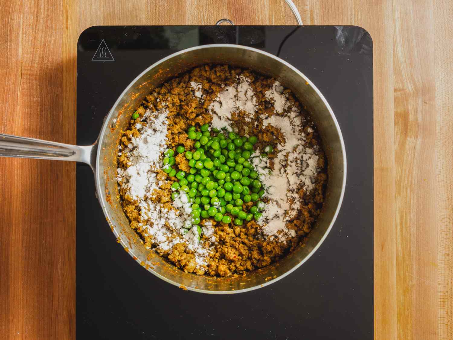 Overhead photo of peas and flour added on top of lamb kheema mixture in a saucepan.