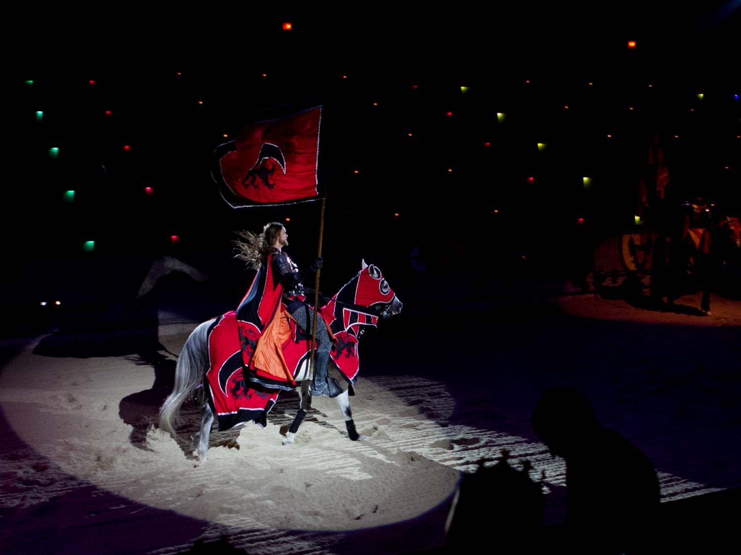 The Red Knight at Medieval Times, on horseback, in full regalia, spotlighted against the sandy floor of the arena