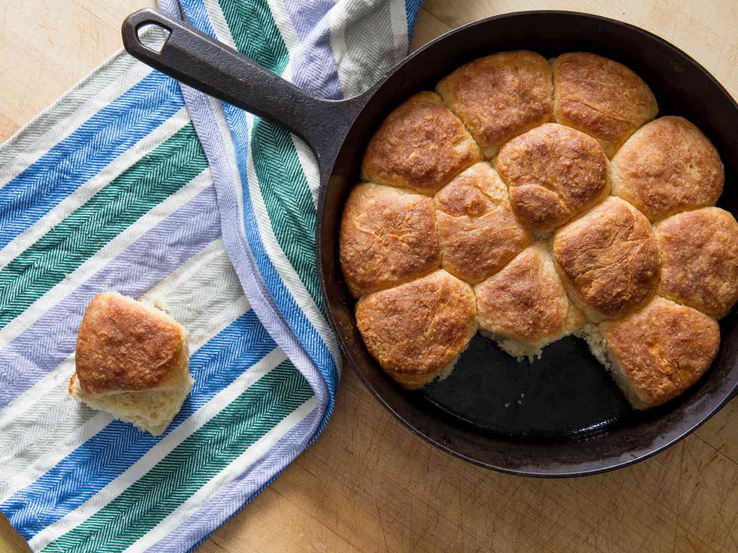 A cast iron skillet of golden brown yeast-raised angel biscuits beside a blue and green striped dishcloth