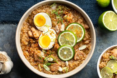 An overhead photo arroz caldo (Filipino chicken and rice soup), topped with a split boiled egg, lime wheels, and sliced scallions. A bulb of garlic and a halved lime is next to the bowl.