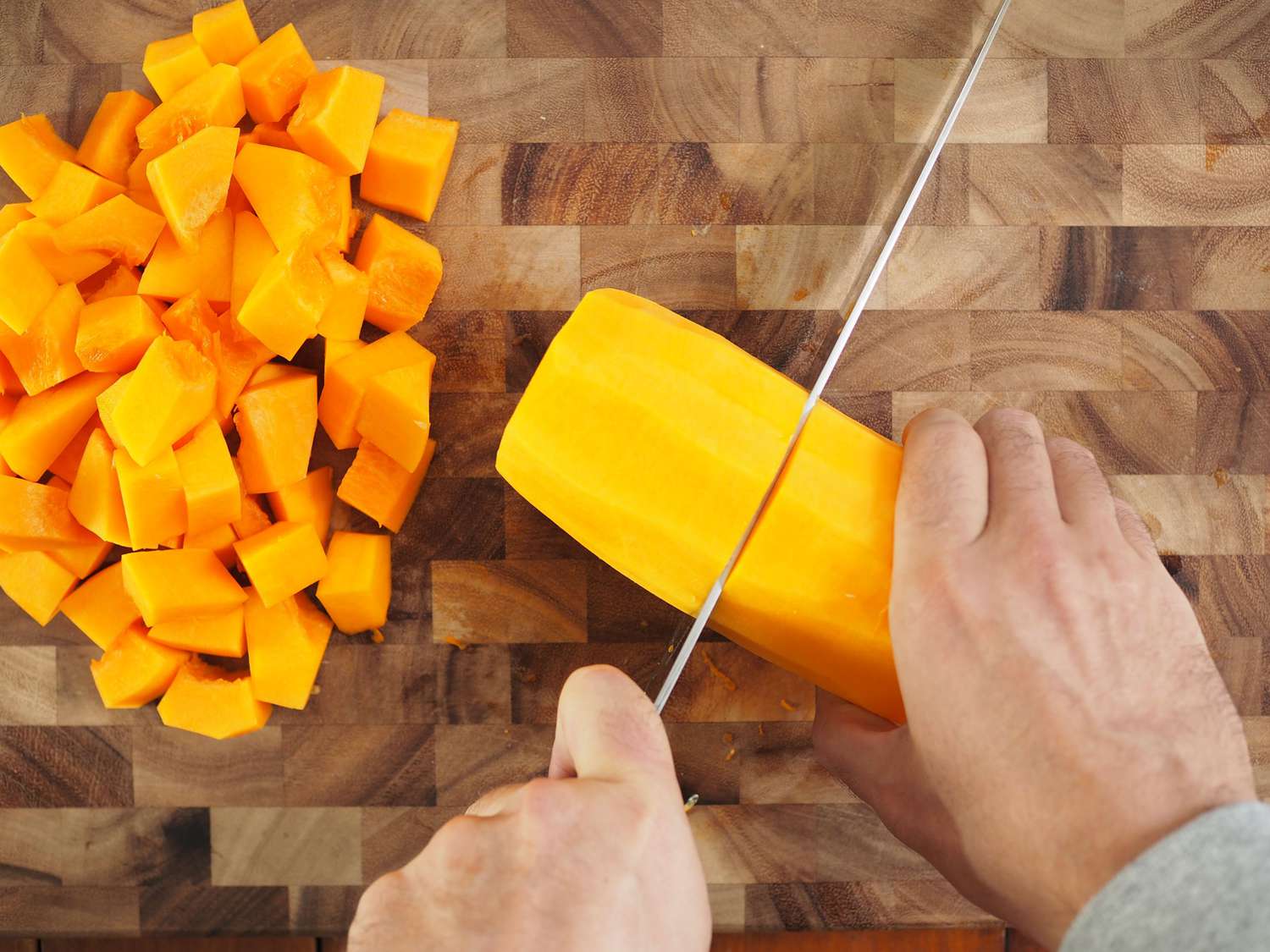 A knife slicing into the neck of a butternut squash. There is a pile of squash chunks on the left side of the image.