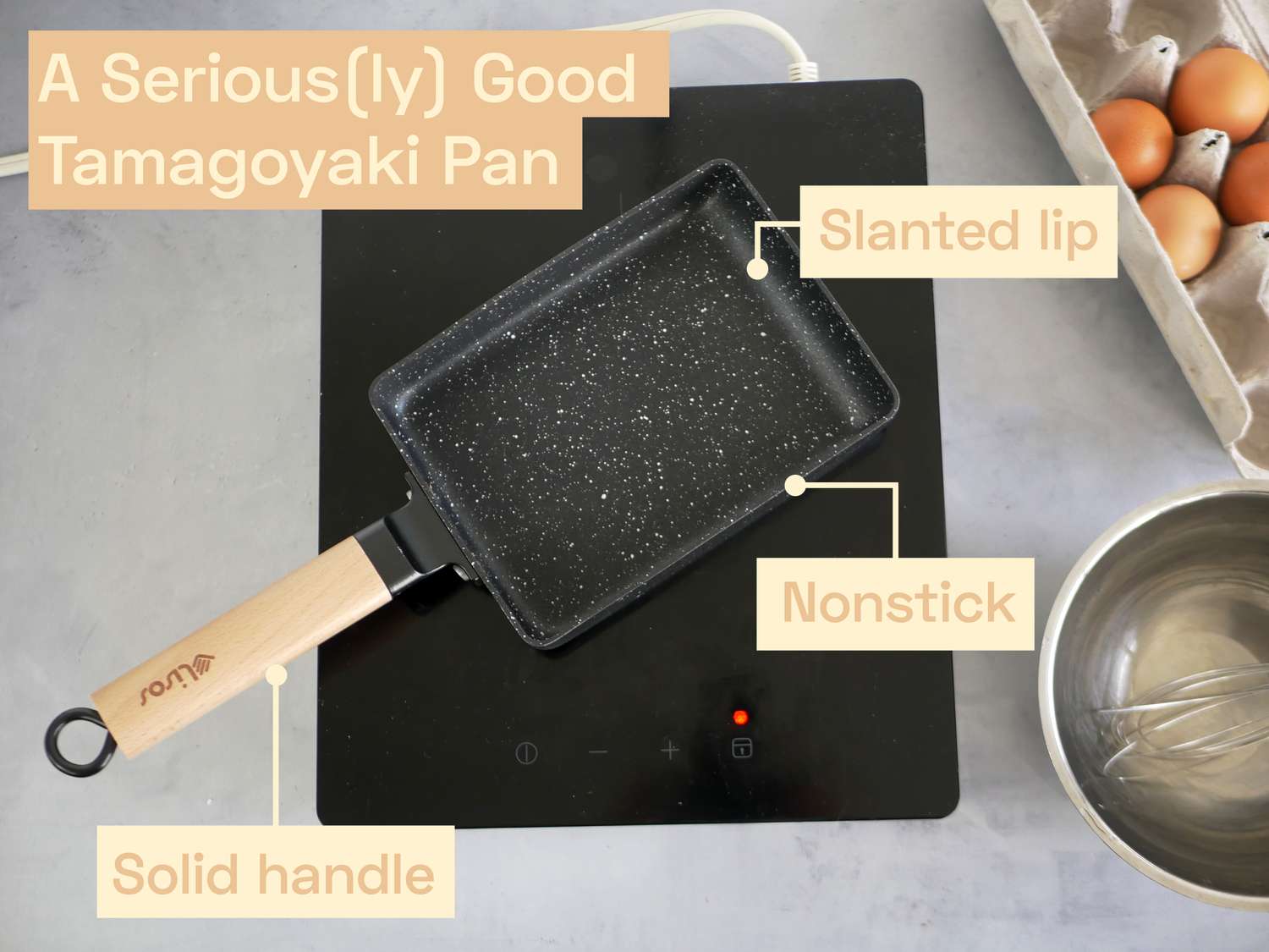 a tamagoyaki pan on an induction burner with a carton of eggs and a meta bowl with a whisk on the side