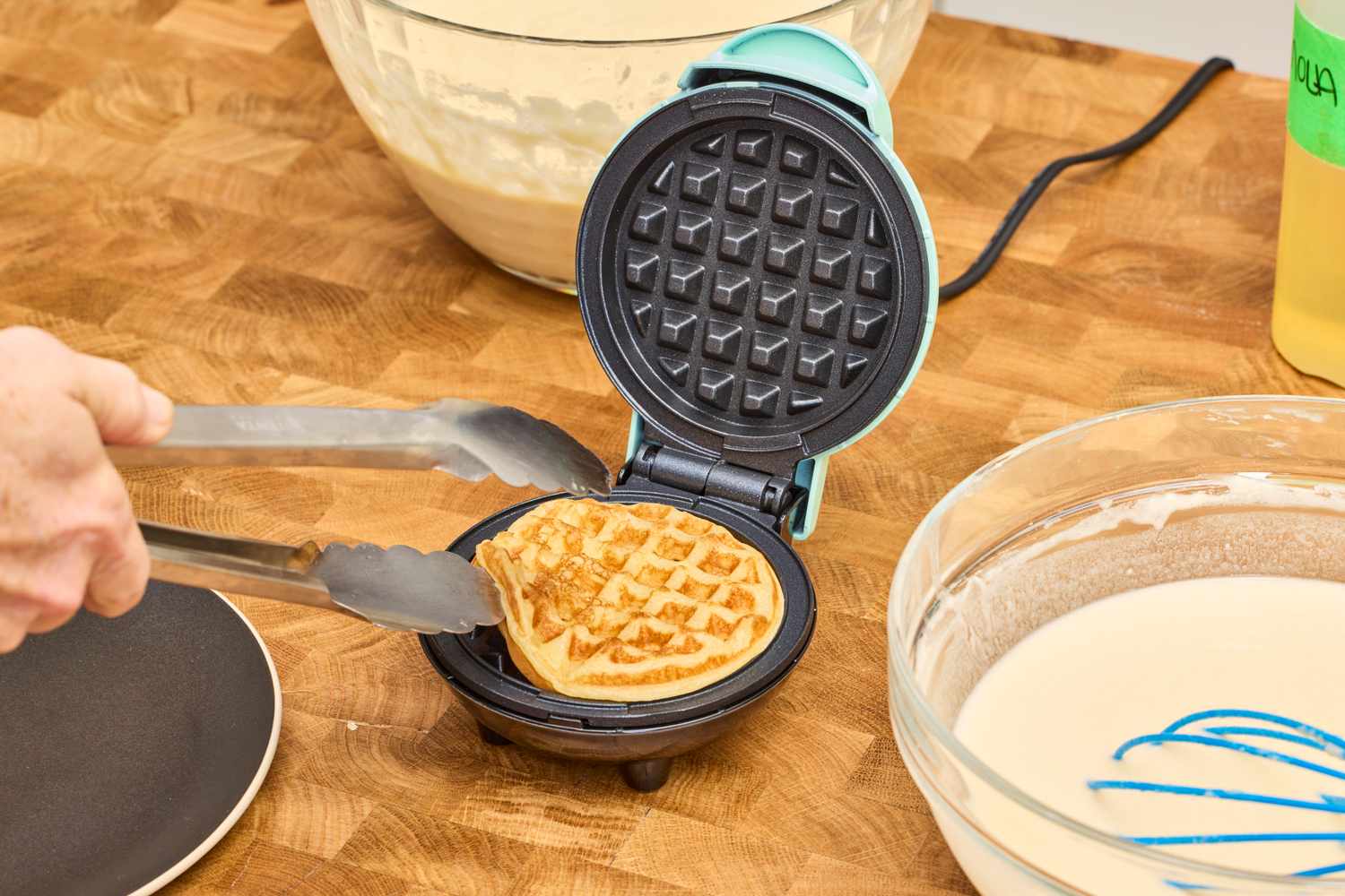 A person using tongs to remove a waffle from the DASH Mini Waffle Maker on a wooden surface next to a batter bowl and dish