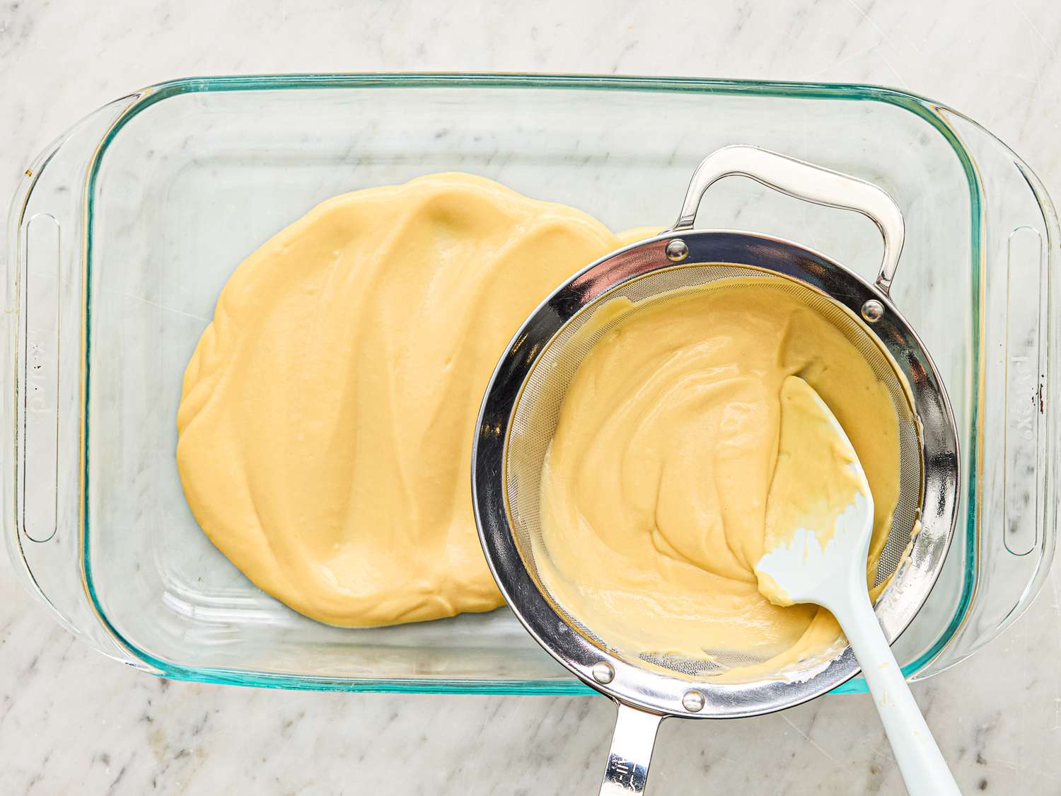 Cream being spread in a glass dish with a spatula resting on a strainer