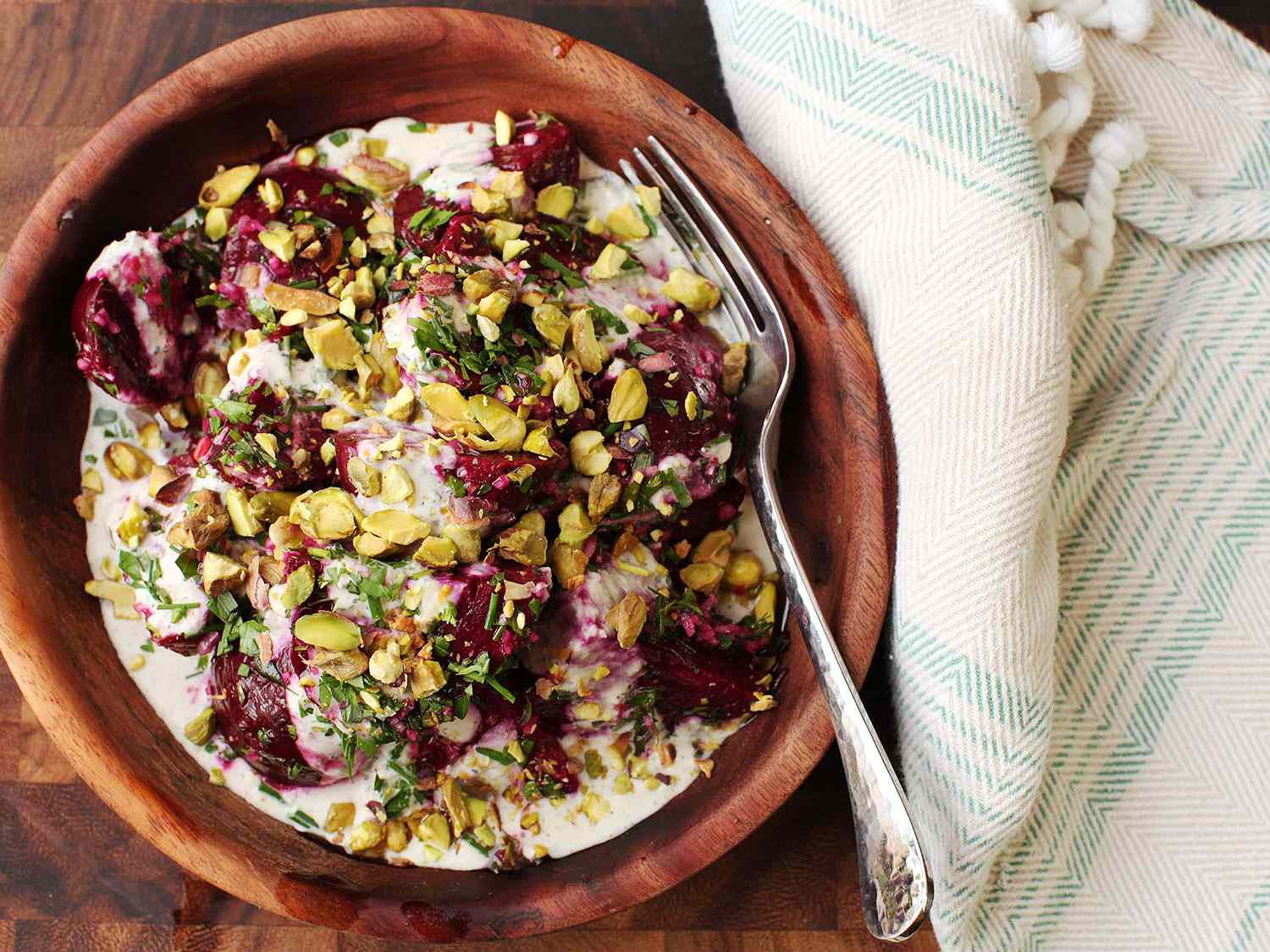 Roasted beet salad with horseradish crème fraîche and pistachios in a wooden bowl with a fork, next to a cream-colored napkin with light blue stripes.