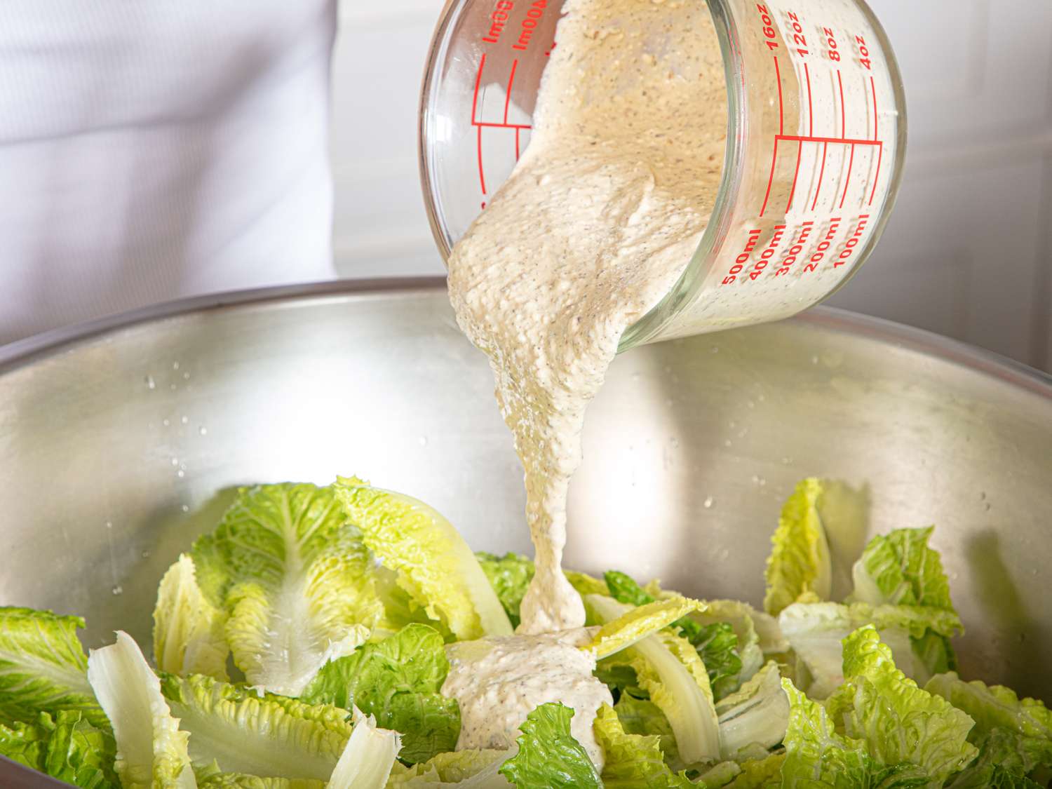 Dressing being poured from a measuring cup onto lettuce in a mixing bowl