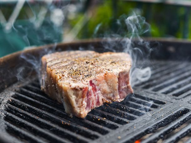 Profile shot of a seasoned porterhouse freshly added to the grill. Wisps of smoke are starting to emanate from the grates.