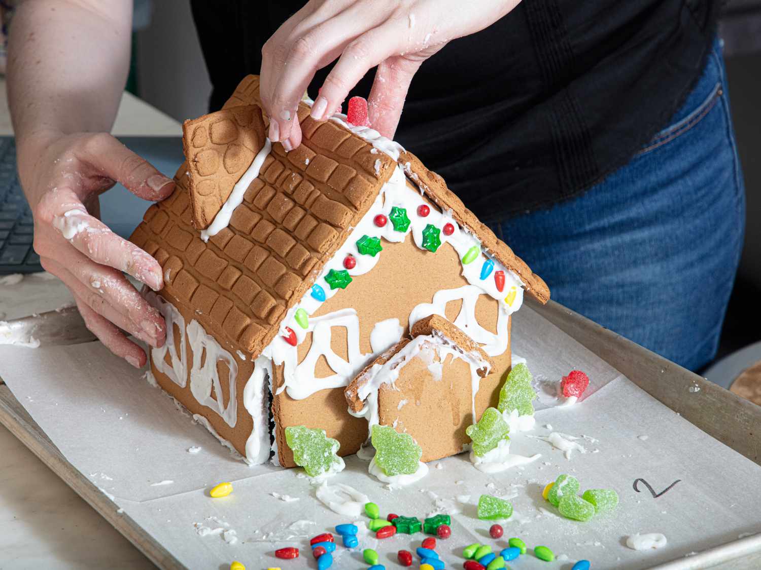 Hands decorating a gingerbread house with frosting and candy on a tray