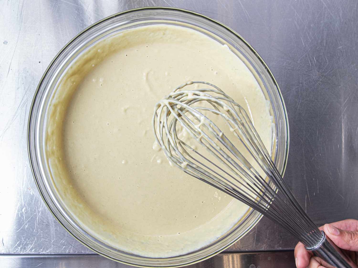Overhead of a whisk dripping over a bowl of mixed tahini and hummus