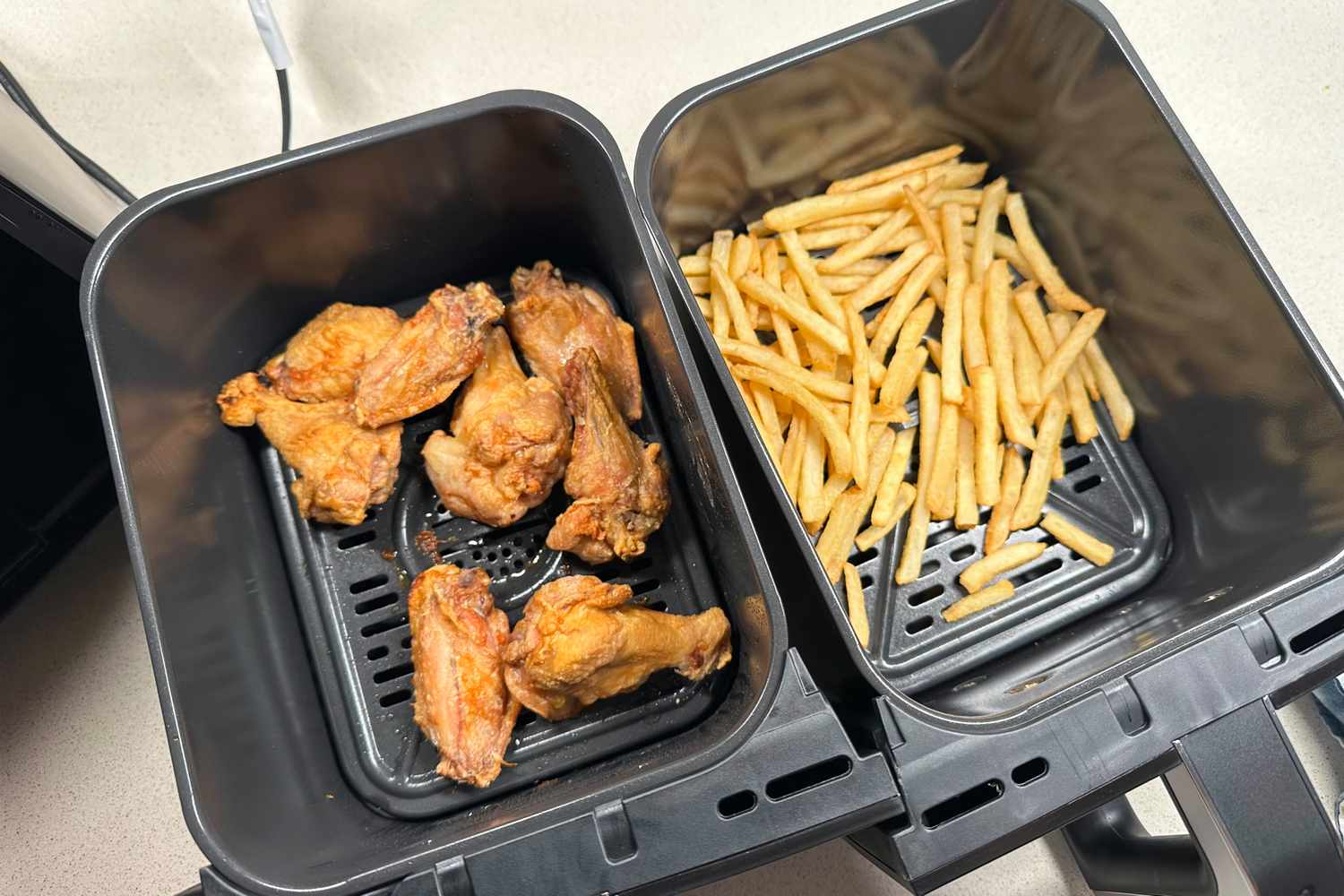 Two baskets of an air fryer one containing chicken wings and the other containing French fries