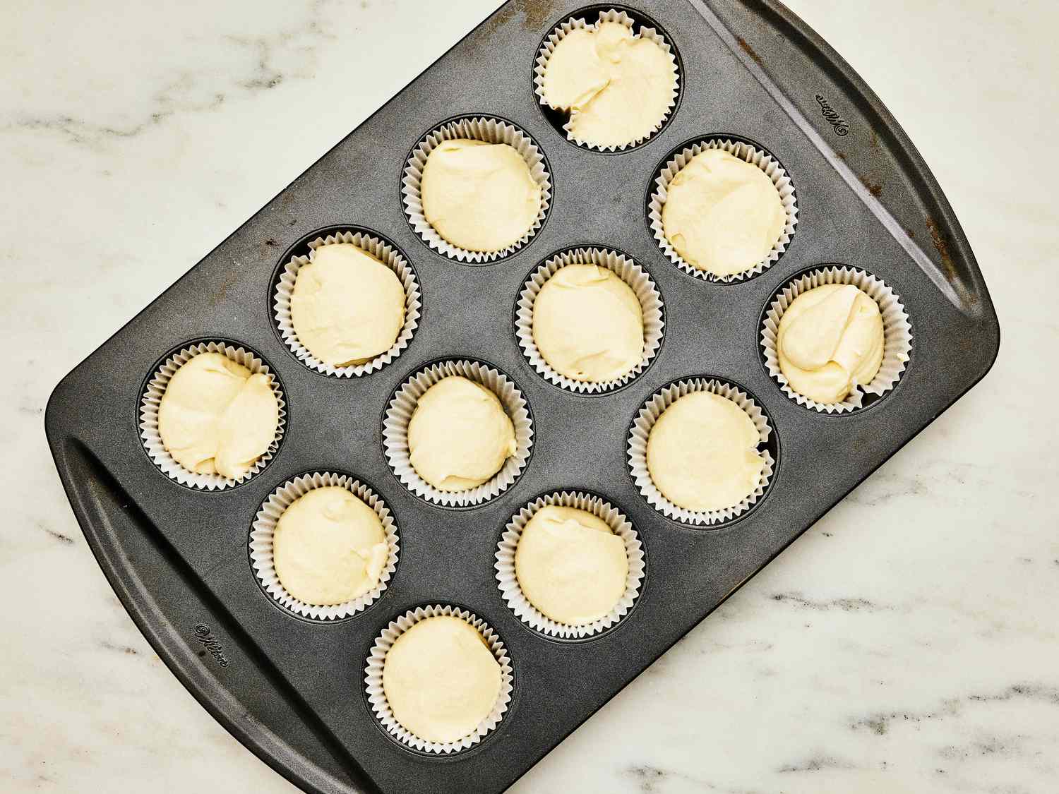 Cupcake batter in baking cups, inside a cupcake tin. The tin is on a white marble tapletop