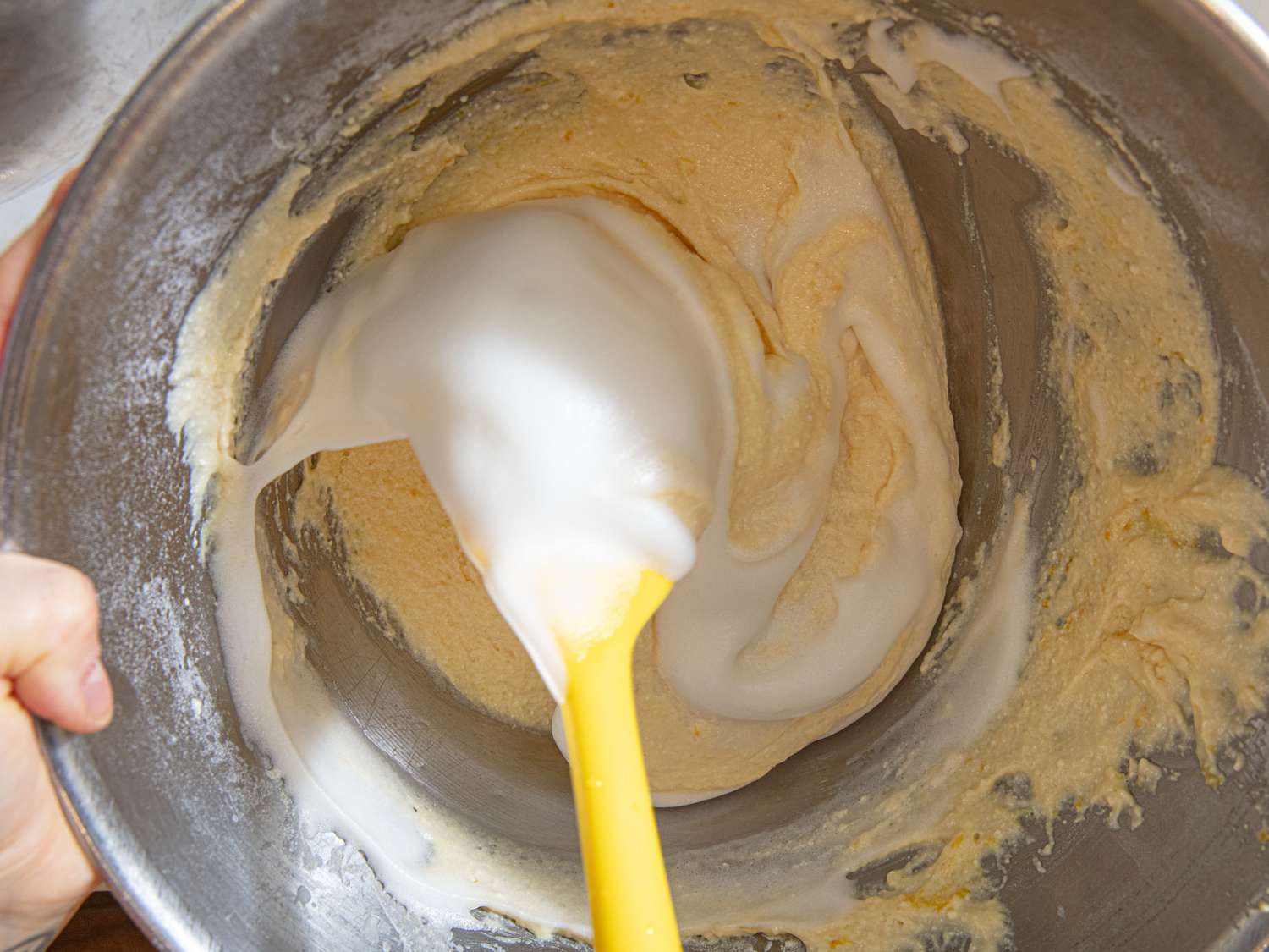 A mixing bowl containing batter being folded with whipped ingredients using a yellow spatula