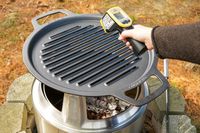 A hand holding a infrared thermometer measuring the temperature of a grill atop a metal outdoor stove with glowing embers beneath