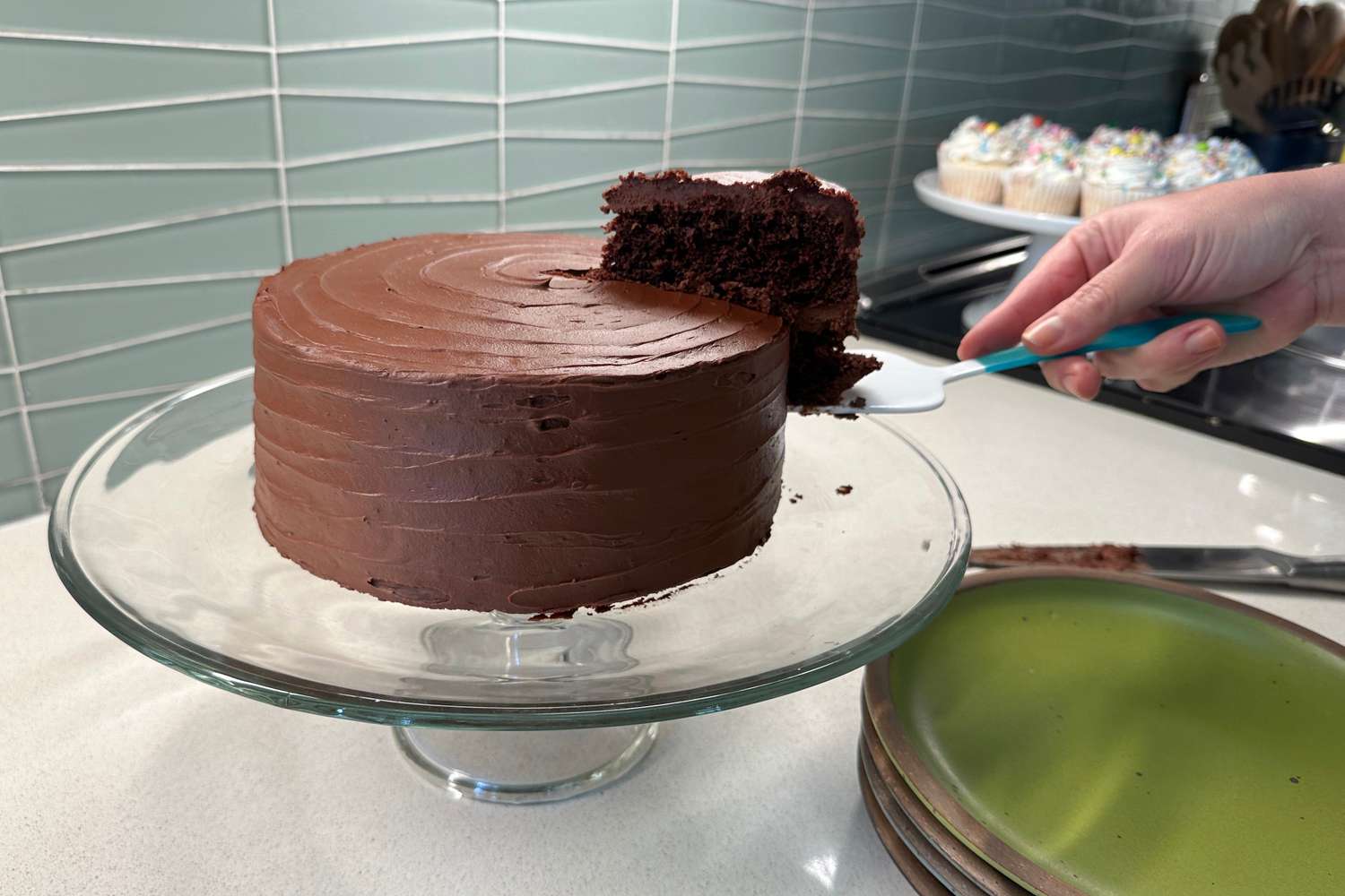 A person lifts a slice of chocolate cake from a glass cake stand
