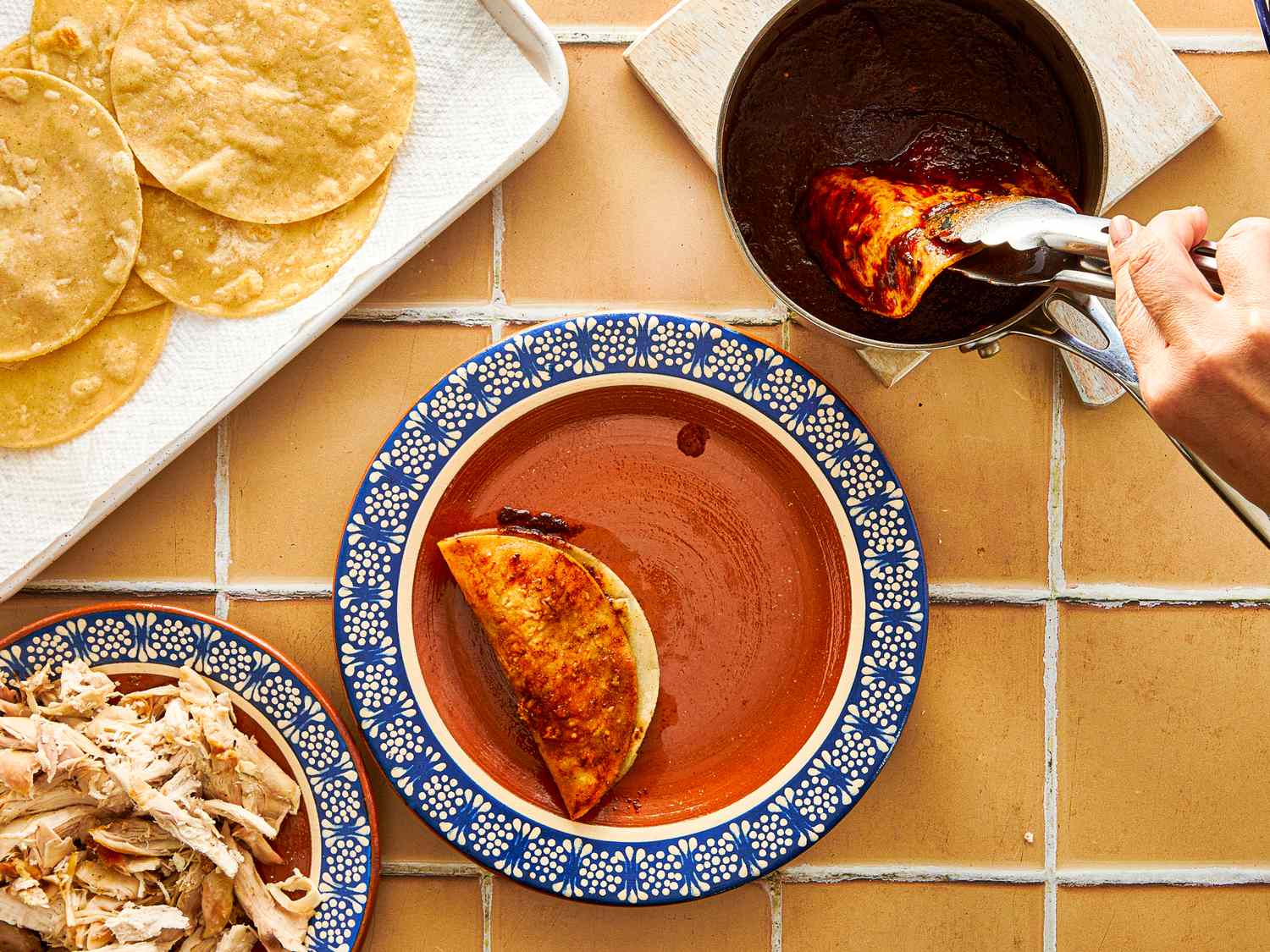 A hand dipping a tortilla filled with chicken into a sauce with tortillas and shredded chicken on the side