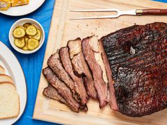 Sliced smoked brisket on a wooden cutting board with pickles and bread on the side