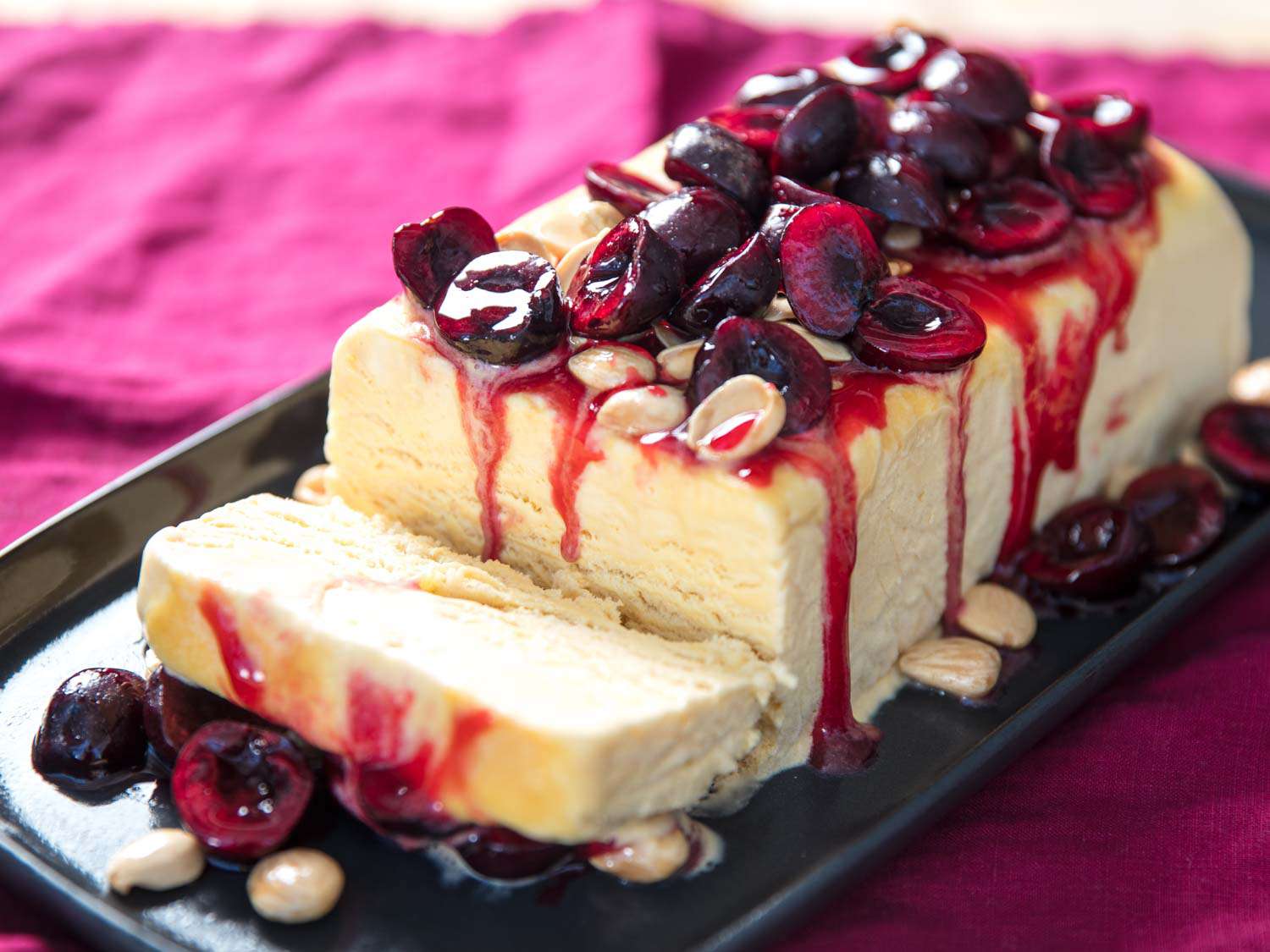 A semifreddo unmolded onto a chilled serving dish, topped with macerated cherries and Marcona almonds. An end slice is leaning toward the camera, propped up by toppings that have fallen to the dish.