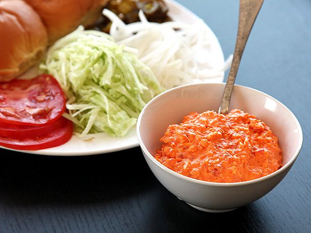 Burger toppings on a plate, including buns, jalapenos, tomatoes, lettuce, and onions, next to a small bowl of pimento cheese with a spoon.