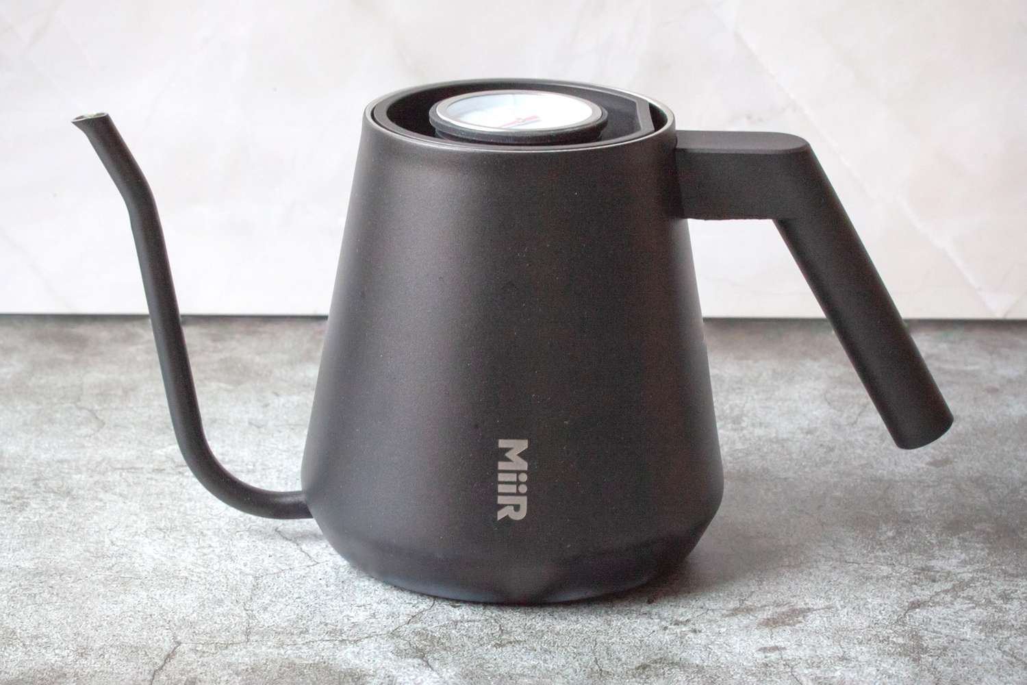 A modern kettle with a spout and a thermometer branded MiiR placed on a textured surface