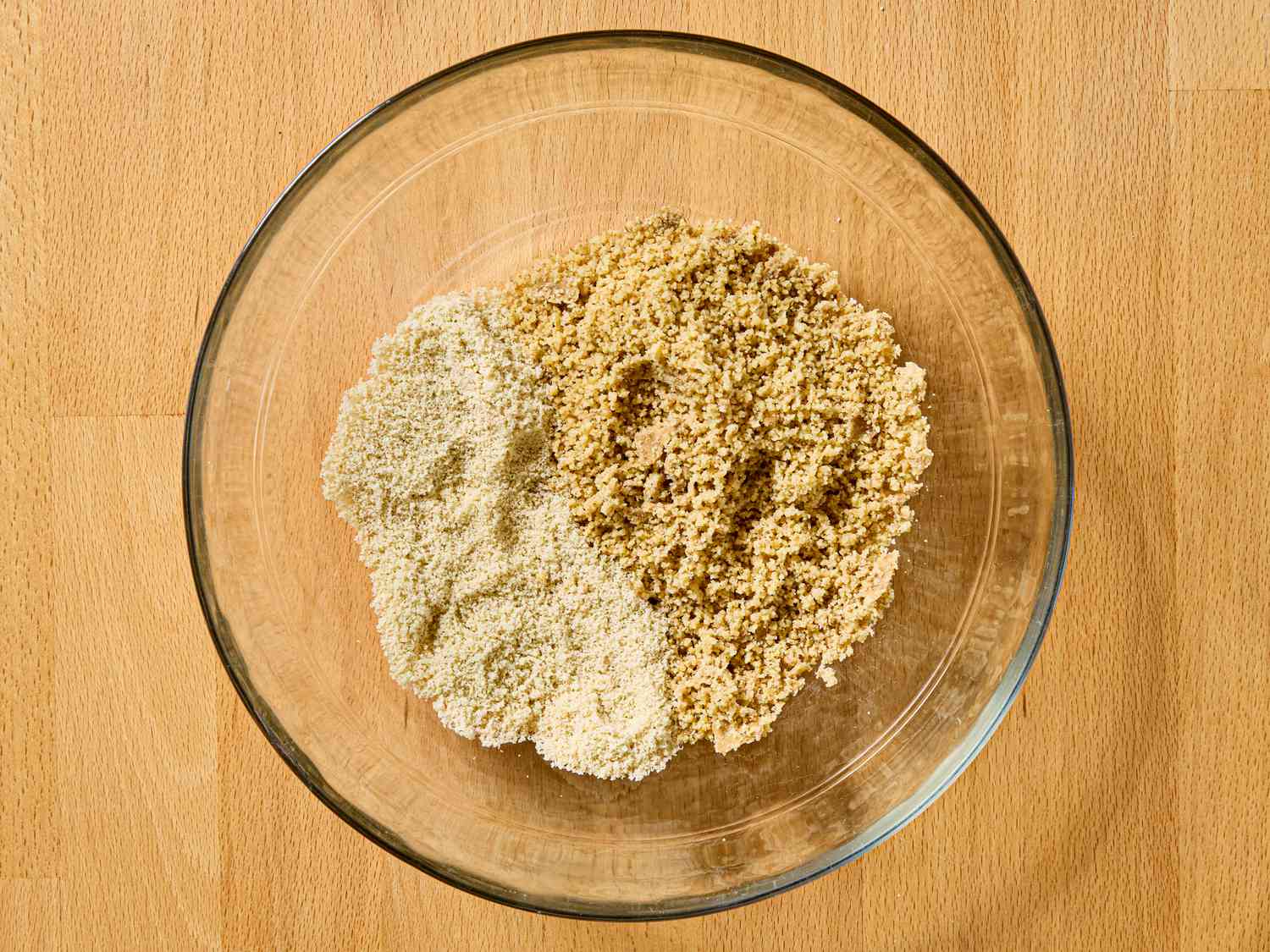 A clear glass bowl containing two types of similar looking ground ingredients placed on a wooden surface