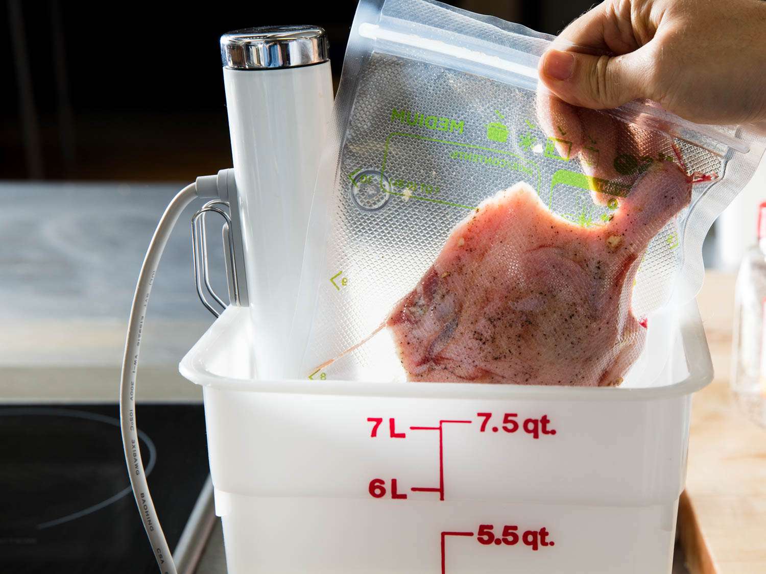 A hand lowering a vacuum-sealed bag of duck leg into a Cambro container with a sous vide circulator attached.