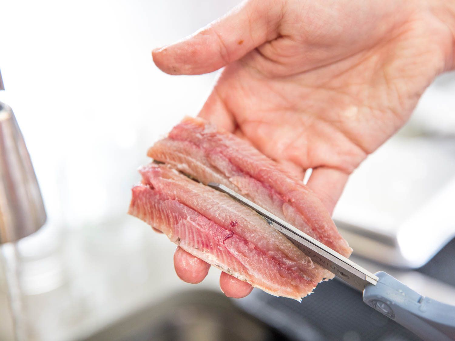 Hands holding a butterflied fresh sardine fillet, and using scissors to cut it into two pieces.