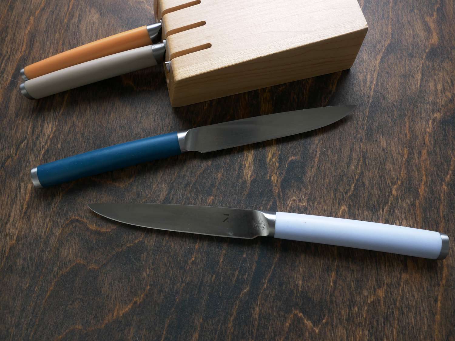 Two material knives on a wooden surface with the knife block with two other knives behind it