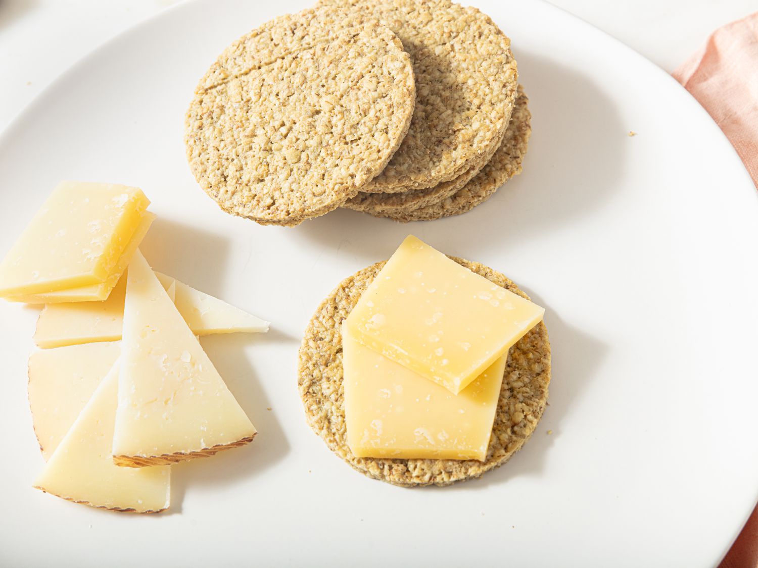 white plate with stack of oatcakes on the top, with slices of cheese to the bottom left and oatcake with cheese on it on the bottom right. 