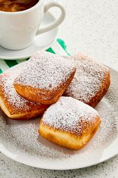 Beignets on a white plate with powdered sugar on it, with a coffee, green and white striped napkin, and a marble surface