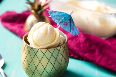 Several scoops of fresh pineapple ice cream, served in a brass-finished chalice with a diamond-etched pattern. The ice cream is garnished with a paper umbrella.