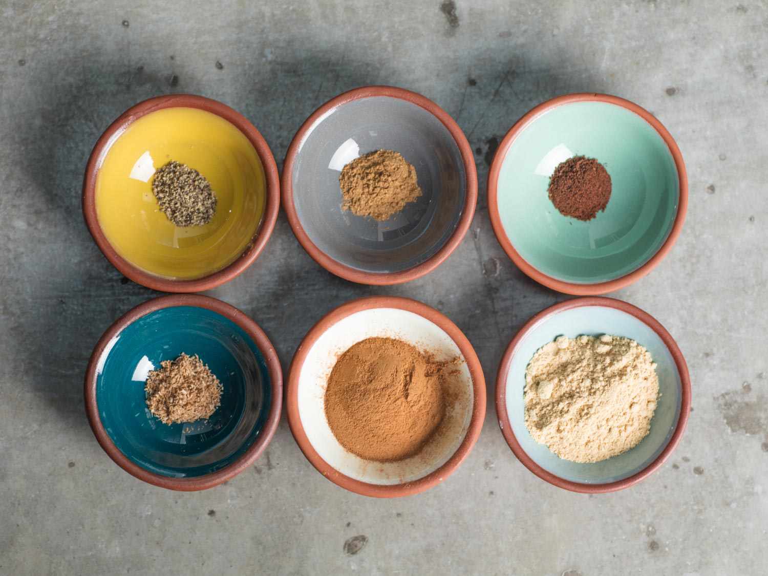 Six small, colorful ceramic prep bowls containing the spices needed for the gingerbread sheet cake.