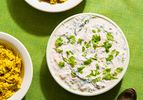 A bowl of raita garnished with herbs next to two bowls of spiced rice and a spoon