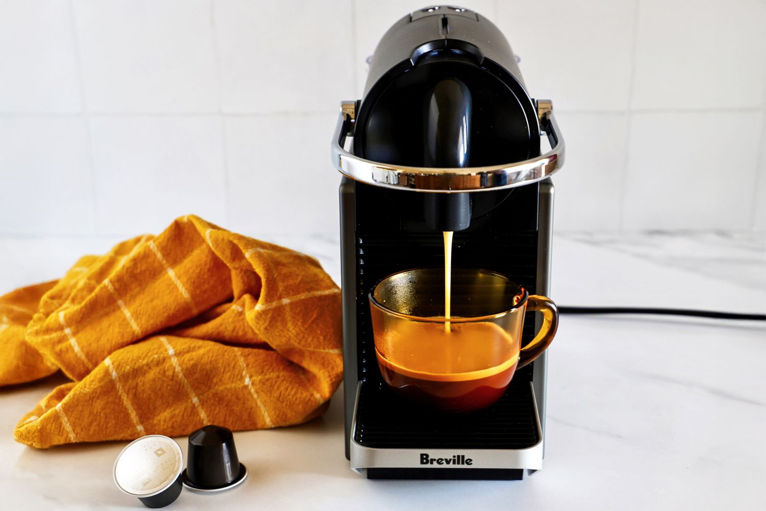 Espresso machine brewing coffee into a glass mug yellow cloth and coffee pods nearby