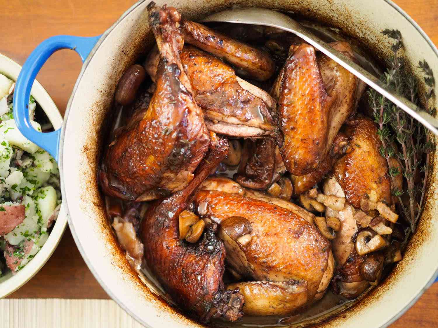 Overhead shot of finished coq au vin on the table, ready to serve from the Dutch oven. A serving bowl of parsley-flecked red potatoes flank the chicken.